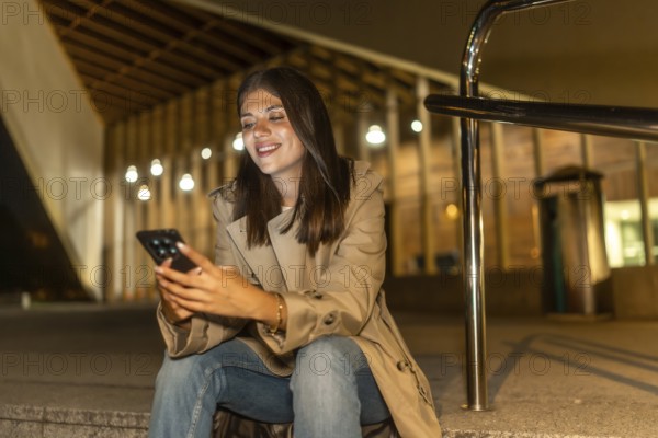 Young woman in a trench coat smiling, sitting on steps in an urban environment at night, connecting with her smartphone and interacting with social media applications