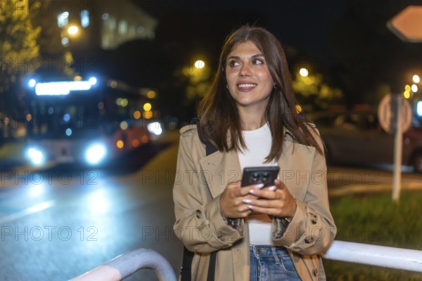 Young woman smiling while checking her mobile phone at a city bus stop at night, anticipating transportation or meeting friends in an urban environment