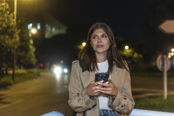 Young woman standing on city street at night, holding smartphone and looking away as warm car headlights and bokeh lights illuminate her contemplative, modern urban moment
