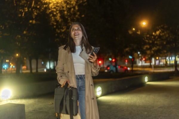 Woman in trench coat walking through an illuminated city park at night, smiling and confident while checking her smartphone, enjoying a peaceful urban evening stroll