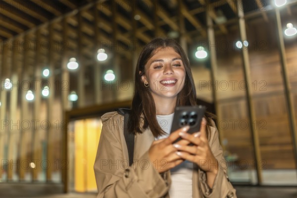 Young woman in a trench coat smiling, checking her smartphone, and experiencing happy moments while navigating modern city life, illuminated by ambient lights and enjoying digital communication