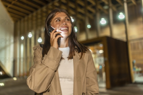 Young woman in trench coat smiling during a phone call, smartphone glow lighting her face on a modern urban street at night with bokeh streetlights and joyful expression