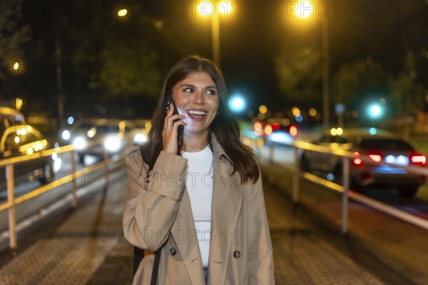 Young adult woman enjoying a happy conversation on smartphone while walking on a city street at night, with blurred streetlights and car lights in the background