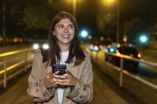 Young woman smiling while holding a smartphone and walking through a downtown street at night, illuminated by bokeh car lights and urban evening glow, casual and confident
