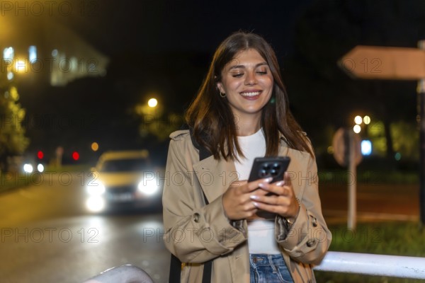 Young brunette woman standing on a city street at night, smiling and checking her smartphone in a trench coat under glowing car lights and bokeh city background