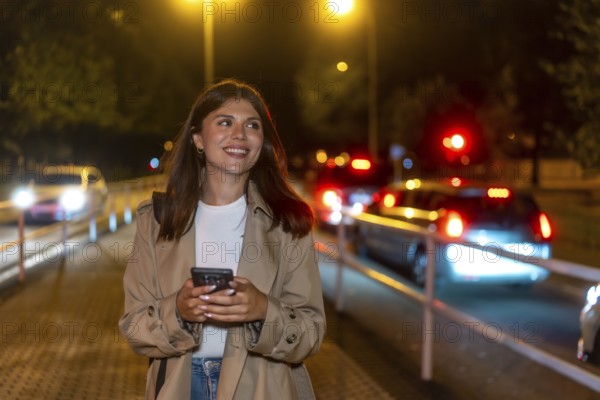 Young woman in a trench coat smiling, holding a phone, and looking away while walking on a city street at night with urban lights and blurred traffic in the background