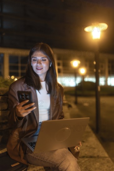 Young woman engaging in remote work and communication, looking at her smartphone while holding a laptop under the glow of urban street lights in a city environment