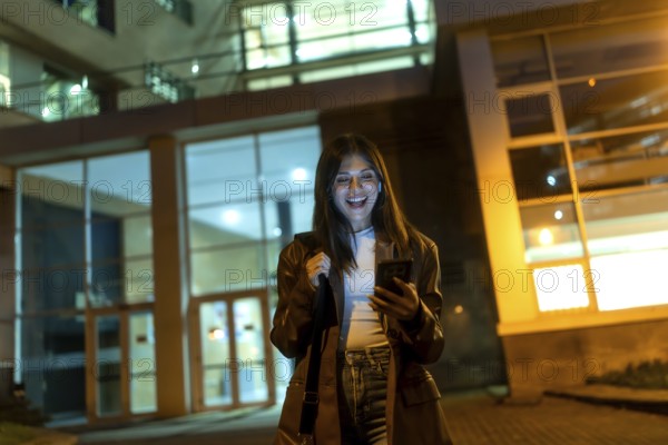 Young woman standing on an urban street at night, happily looking at her smartphone while holding a shoulder bag, with a modern building's illuminated facade in the background