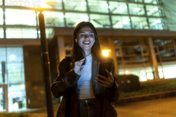 Young woman smiling broadly, wearing wireless earbuds and dark jacket, interacting with a smartphone on a city street at night, with illuminated buildings in the background