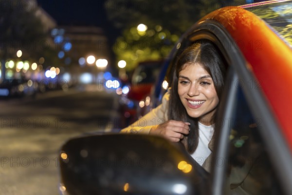 Young woman leaning out of a car window at night, smiling as city lights and bokeh blur by, enjoying a joyful, free evening ride through urban streets