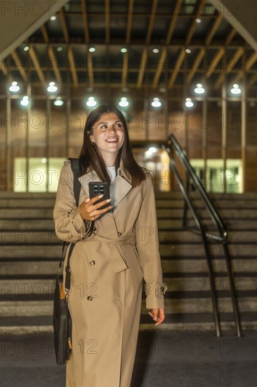 Young woman dressed in a trench coat smiling, holding a mobile phone, and walking up illuminated stairs in a modern building at night after work, showing urban lifestyle