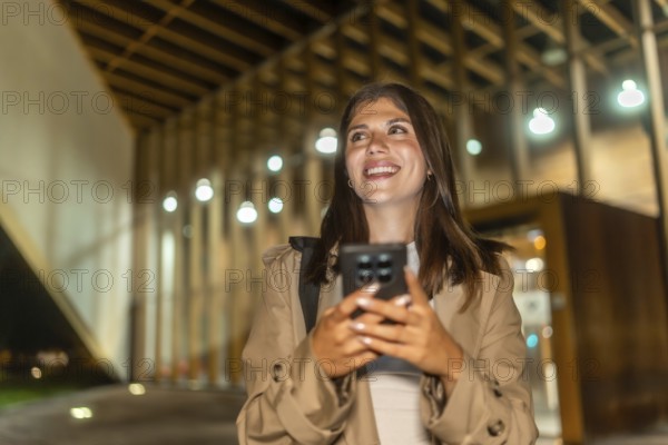 Young woman smiling, holding phone while walking illuminated city streets at night, confidently navigating with an app, feeling connected, relaxed and modern in urban lifestyle scene