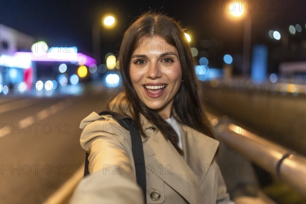 Young woman smiling and taking a selfie on a lively city night, bokeh street lights and light trails glowing behind her as she explores urban streets in a trench coat