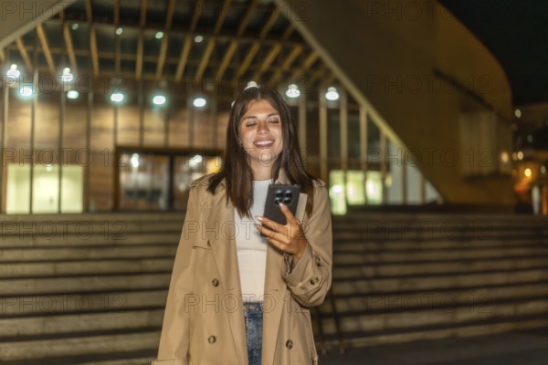 Young woman smiling while looking at her mobile phone outdoors, standing at night in front of a modern building with illuminated steps, connecting with technology and enjoying urban life