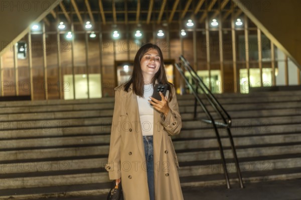 Young woman in a trench coat smiling and looking at her smartphone while standing on modern urban stairs at night, connecting with others or navigating the city