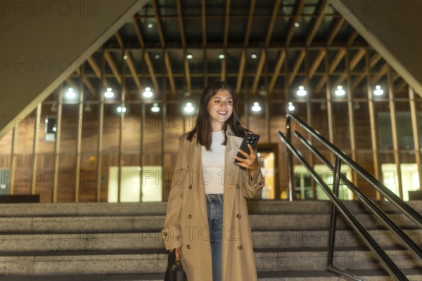 Young woman smiling while checking her smartphone, walking down urban stairs at night, wearing a trench coat, reflecting modern communication and city life