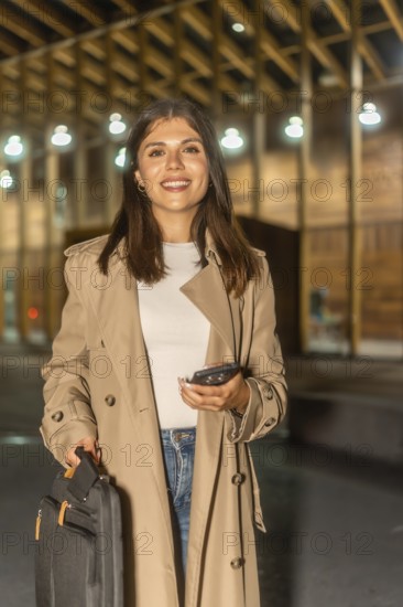 Young businesswoman traveling and commuting, holding a smartphone and briefcase, smiling while walking at night in an urban city environment, representing business travel