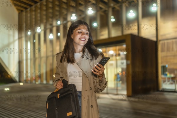 Young businesswoman smiling while checking her smartphone, holding a briefcase and walking alone on an illuminated urban street after work, embodying modern professional life and connectivity