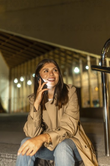 Young woman sitting outdoors at night, smiling brightly while having a phone conversation, illuminated by the smartphone screen in an urban environment