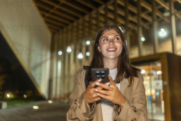 Young woman smiling while holding a smartphone and looking up, exploring a modern city street at night, experiencing technology and connectivity in an urban setting