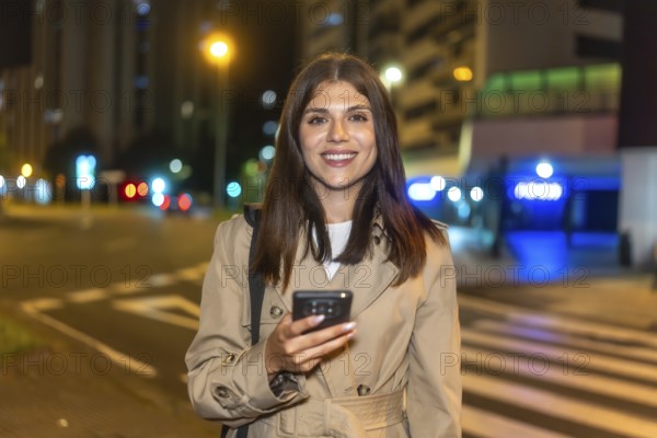 Woman smiling brightly and holding a smartphone, standing on a bustling city street at night with illuminated buildings and traffic lights creating a vibrant urban background