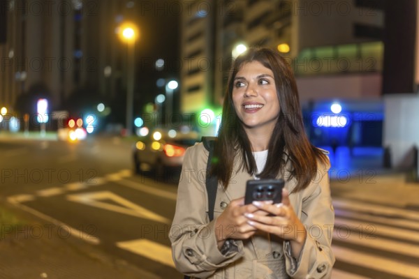 Young woman smiling and looking away while using a mobile phone on a pedestrian crossing, waiting for a ride or navigation in a modern illuminated urban environment after sunset