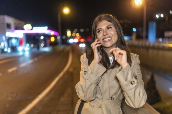 Young woman in a trench coat smiling as she talks on her mobile phone on a bustling city street at night, bathed in warm urban lights and soft bokeh background