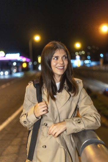 Woman smiling on a nighttime city street, wearing a trench coat, looking away as colorful urban lights and bokeh create a vibrant, stylish, cheerful scene of evening exploration