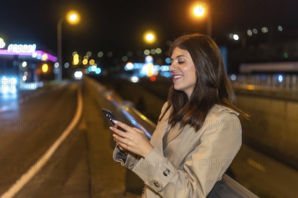 Young woman standing on an urban bridge at night, smiling while interacting with her smartphone, with blurred city lights and a road creating a vibrant bokeh background