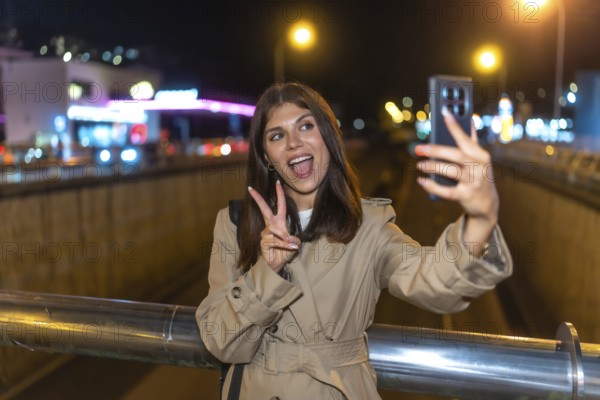 Young woman wearing a trench coat standing on a bridge, holding a smartphone and taking a self portrait, showing the peace sign with a happy expression during a vibrant city night