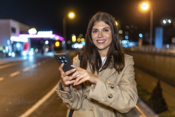 Young woman in a trench coat standing on a city street at night, smiling at her smartphone as she texts and browses, enjoying urban connectivity and glowing lights around her