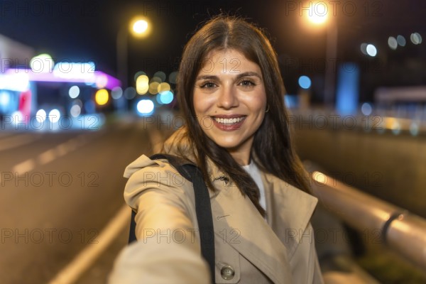 Young woman smiling and holding a smartphone, capturing a selfie portrait outdoors in an urban environment at night, surrounded by blurred city lights and a vibrant atmosphere