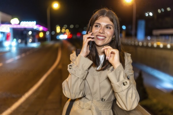 Young woman in a trench coat smiling happily while having a phone call, standing on a city street at night with blurred lights creating a vibrant urban background