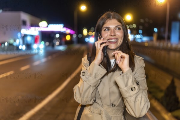 Young woman in a trench coat smiling and laughing while talking on her smartphone during an illuminated city night stroll, enjoying a relaxed, joyful conversation