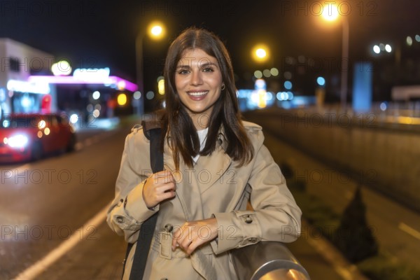 Young woman smiling while standing in a trench coat on an urban street at night, illuminated by city lights and a gas station in the blurred background