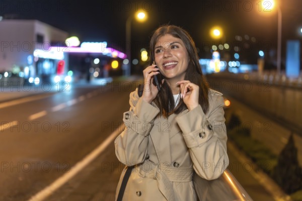 Young woman smiling and communicating on a smartphone, walking along a vibrant urban street during nighttime with city lights glowing in the background