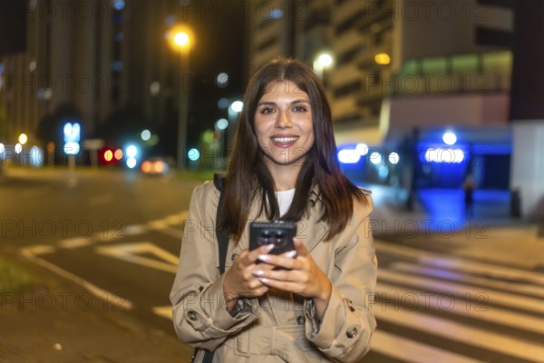 Young woman smiling and interacting with her smartphone while standing on a city street at night, with blurred lights and urban infrastructure providing a dynamic background
