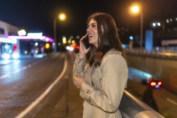 Happy young woman smiling and talking on a mobile phone while standing on an urban street at night, city lights and traffic bokeh creating a modern, lively backdrop
