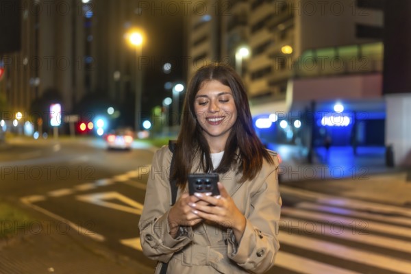 Young woman smiling with eyes closed, enjoying her smartphone on a city street at night, relaxed and content while texting, browsing social media amid glowing streetlights and bokeh