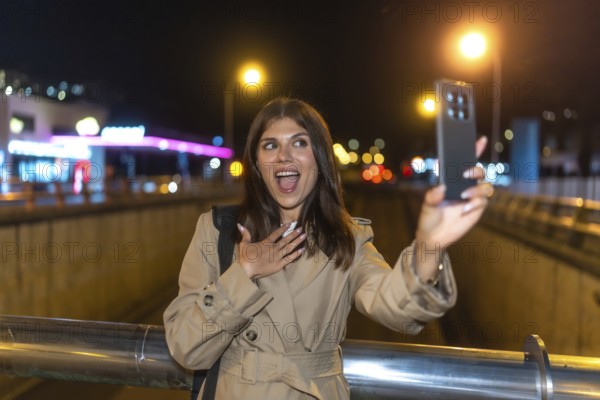 Young woman in trench coat surprised and joyful taking a nighttime selfie on a bustling city street, phone glowing against blurred lights for vlogging and social sharing