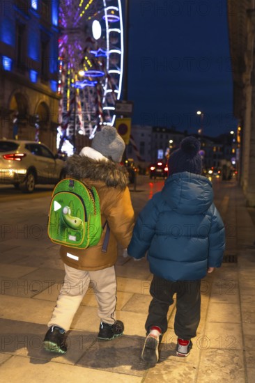 Two children holding hands and walking away from the camera on a sidewalk near a brightly lit ferris wheel at christmas time in bayonne, france