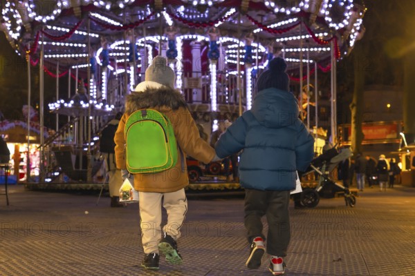 Two young brothers, holding hands, walk away from a brightly lit carousel at a christmas market in bayonne, france, enjoying the festive atmosphere