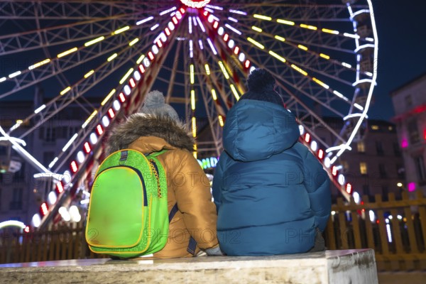 Two children with winter coats and backpacks admiring a brightly lit ferris wheel at a christmas market in bayonne, france