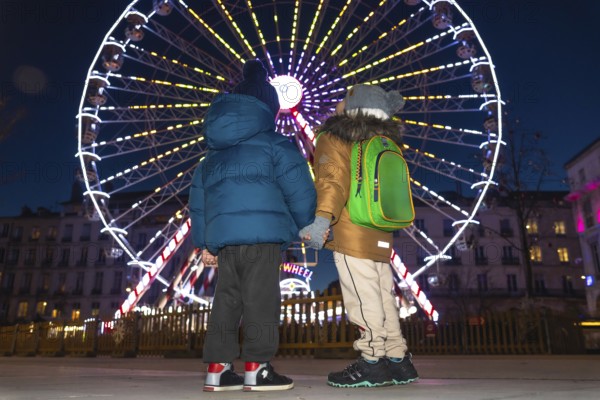 Two children holding hands are admiring a brightly lit ferris wheel at a christmas market in bayonne, france, creating a magical winter holiday scene