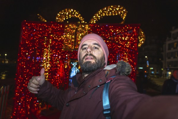 Tourist taking a selfie and making thumbs up sign with christmas lights and decorations in bayonne, france