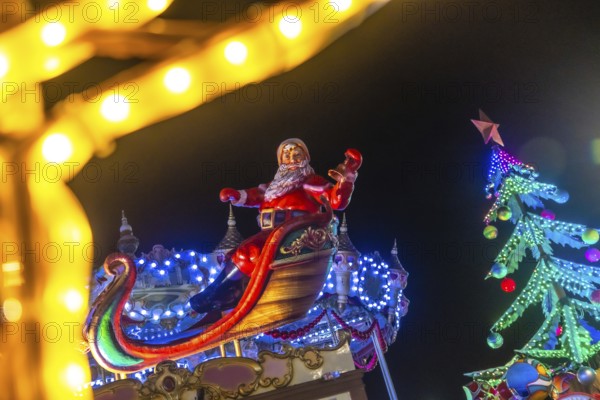 Santa claus sitting on his sleigh and waving to people during christmas night in bayonne, france, with a colorful christmas tree and lights