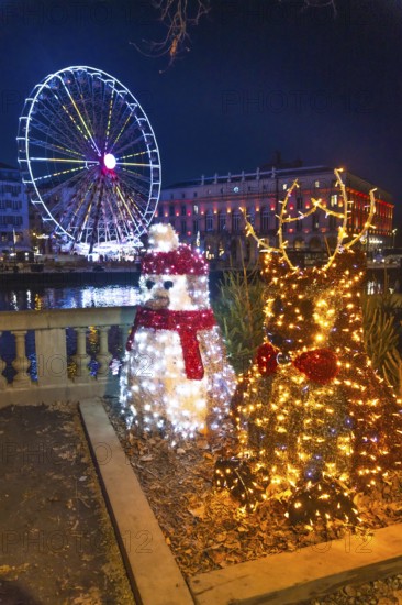 Luminous christmas decorations featuring a reindeer and a penguin stand by the nive river with a brightly lit ferris wheel and bayonne city hall in the background