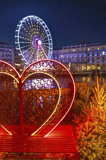 Illuminated heart shaped bench with small christmas trees and ferris wheel in the background, creating a romantic atmosphere in bayonne, france
