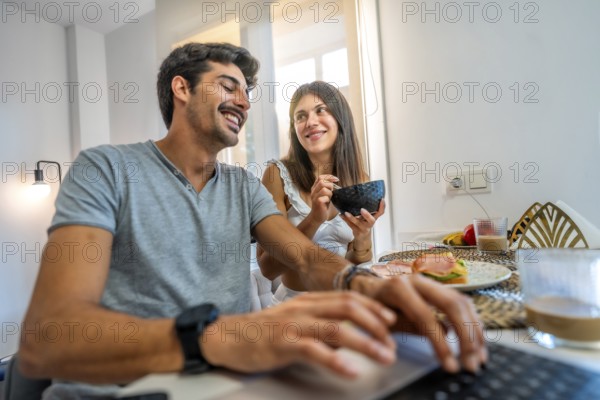 Young couple enjoying breakfast in a bright kitchen, working from home on a laptop while sharing smiles and conversation together