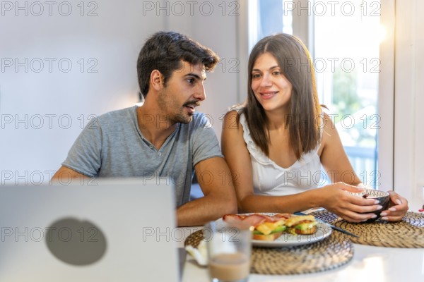 Young couple talking and having breakfast in their kitchen, with a laptop on the table and natural light coming from the window